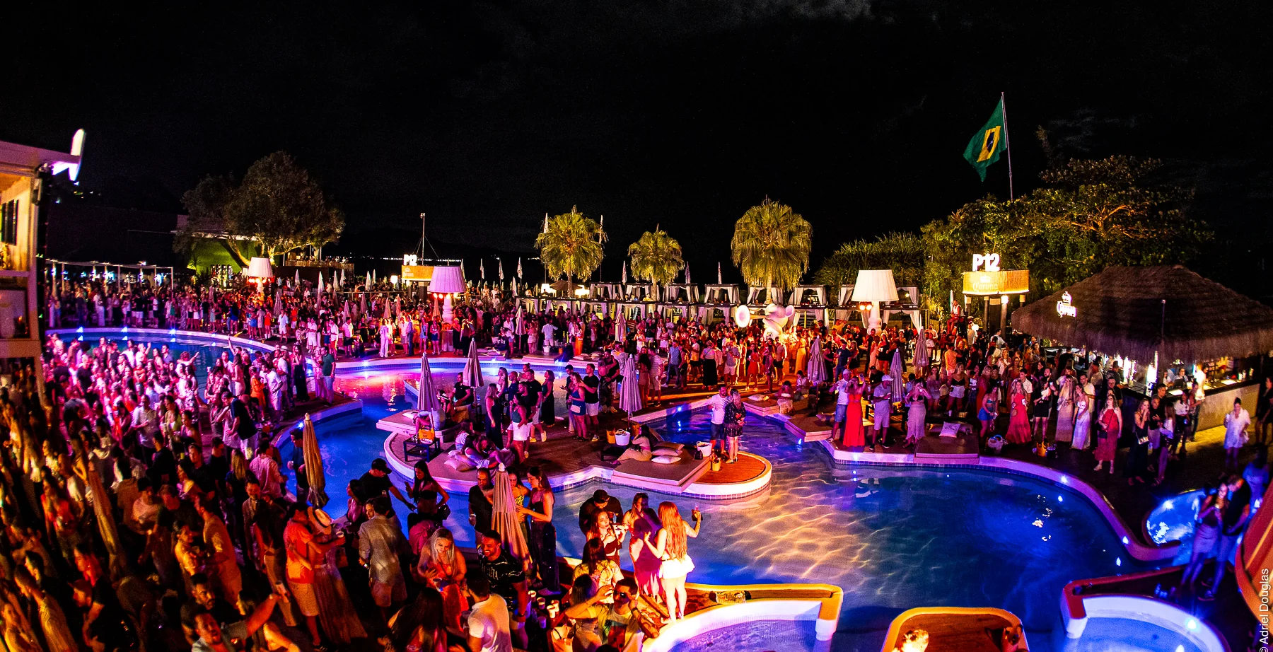 O P12 lotado à noite: a multidão em volta das piscinas iluminadas, as palmeiras e a bandeira do Brasil