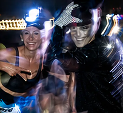 Personagem de luva brilhante e rosto pintado posando com uma atleta, entre luzes em movimento, numa edição anterior