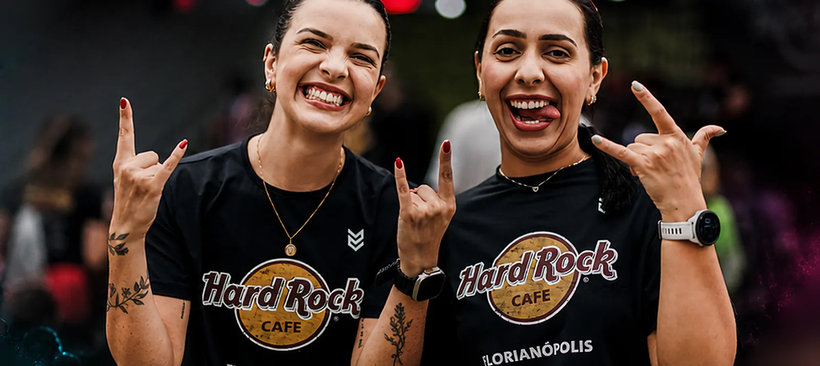 Duas atletas sorrindo e fazendo o sinal do rock com as mãos, de camiseta preta do Hard Rock Cafe Florianópolis