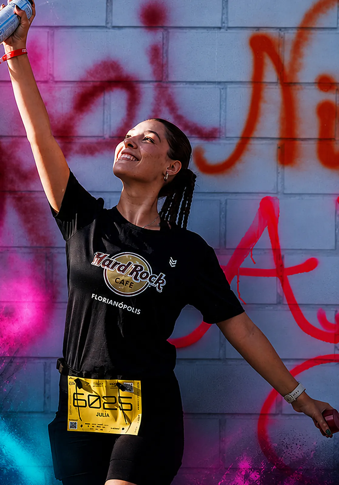 Atleta sorridente ergue uma lata de spray diante de um muro grafitado em magenta e vermelho, com a camiseta do Hard Rock Cafe Florianópolis e o número de peito