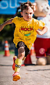 Menino correndo com a camiseta amarela da Corrida Rock Kids, mascote urso ao fundo