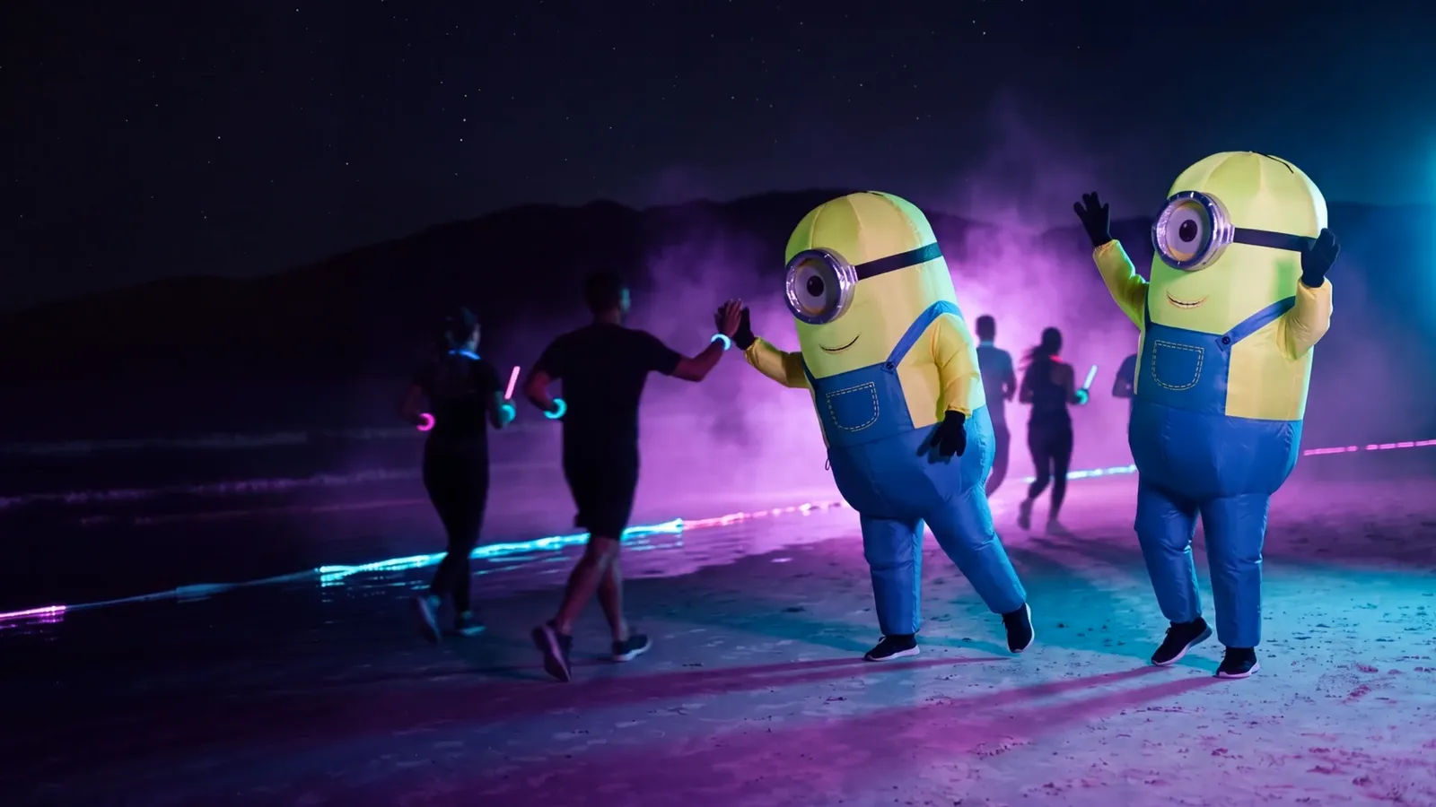 Dois performers em fantasias infláveis amarelas de um olho só batendo na mão de atletas na areia de Jurerê à noite (imagem ilustrativa)