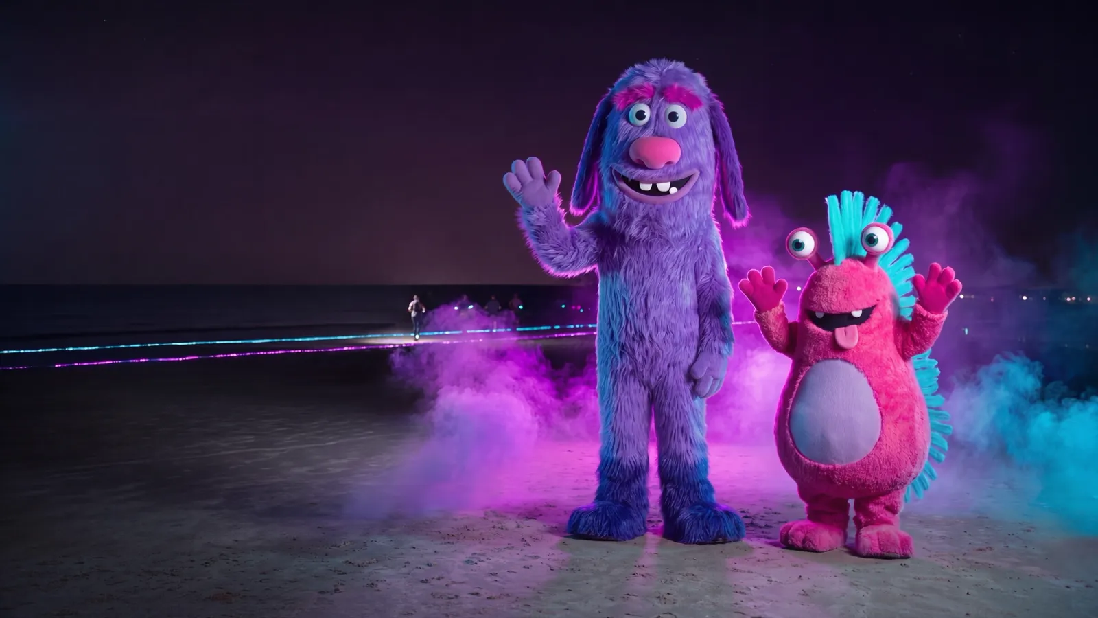 Dois performers em fantasias gigantes de monstro, um alto, violeta e peludo e outro baixinho e magenta com olhos em antenas, acenando para atletas na areia (imagem ilustrativa)